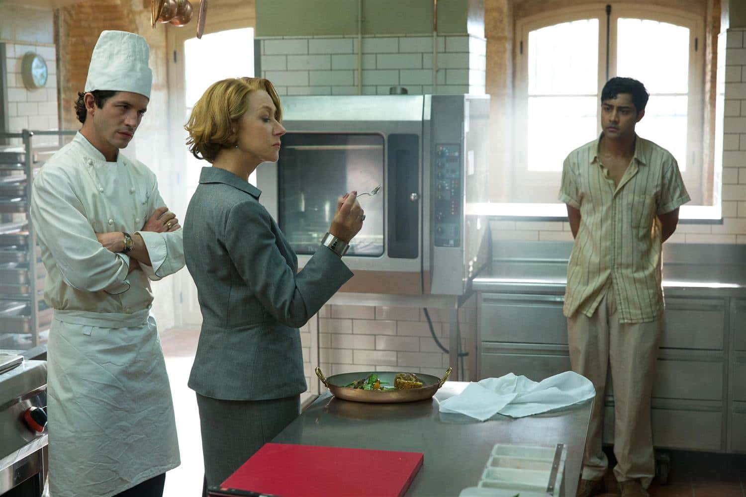 Från filmen 100 steg från Bombay till Paris. En kvinna står i ett kök och provsmakar mat medan två kockar ser på. 
